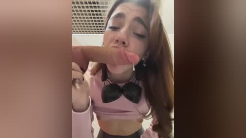 A video of a young, light-skinned woman with long brown hair, wearing a pink crop top with a black bow, sucking a large, erect penis. Background includes a ceiling with square tiles.