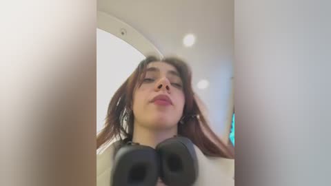 Video of a young woman with long, wavy brown hair and light skin, wearing black headphones, looking upward. She is in a modern, well-lit interior with white walls and recessed ceiling lights.