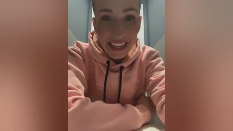 Media: Video of a smiling, fair-skinned, bald man with a trimmed beard, wearing a pink hoodie, leaning against a light blue wall.