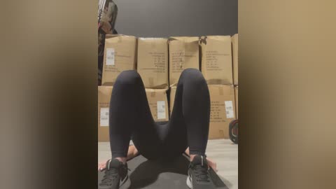 Media: A video of a person in black leggings and athletic shoes, lying on their back in a warehouse with stacks of cardboard boxes in the background.