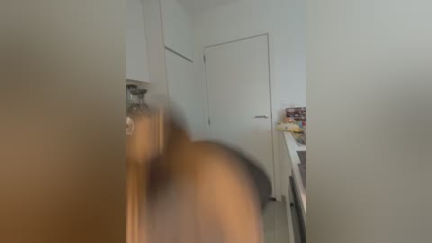 Media: A blurry video of a person with a light skin tone, likely a woman, walking down a hallway. The background features white walls, a door, and a kitchen with visible cabinets and items on a counter.