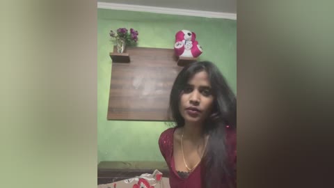 Media: Video of a woman with long black hair and medium brown skin, wearing a red blouse, standing in a room with green walls and a wooden shelf holding a red and white shoe and a bouquet.