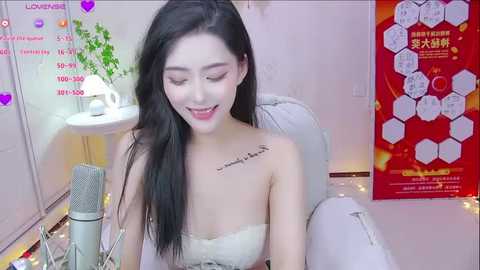 Media: A video of an East Asian woman with long black hair, wearing a strapless white dress, sitting on a grey chair with a microphone. She smiles, and the room has a pink wall with a poster and plants.