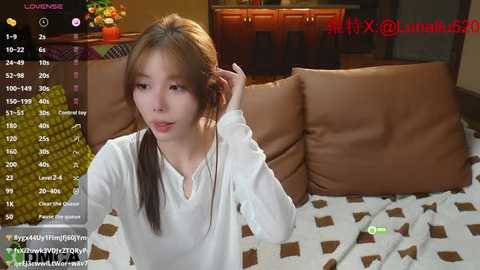 Media: A screenshot of a young Asian woman with light skin and straight brown hair, wearing a white shirt, sitting on a couch. The background includes wooden cabinets and a TV screen displaying a video call.