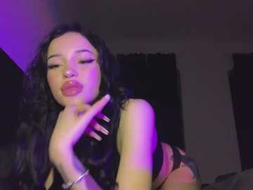 Media: Video of a young woman with long, wavy black hair, wearing a black lace bra, pouting seductively, her right hand touching her lips. The background is dimly lit with purple and blue lighting.