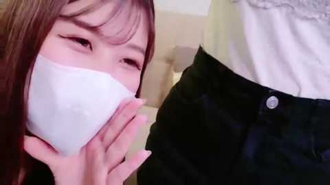 A video of an Asian woman with long brown hair and a white surgical mask covering her mouth, looking concerned, standing beside a person in black pants and a white shirt.