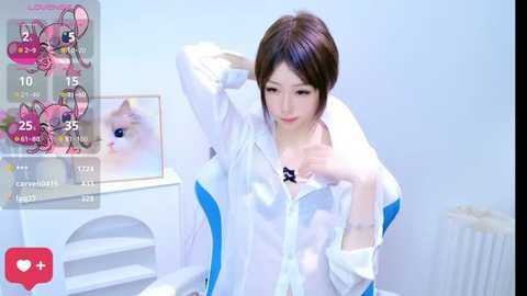 Media: Video of an Asian woman with straight black hair, wearing a white and blue shirt, holding her collar. Background includes a white wall, a radiator, and a framed picture of a cat.