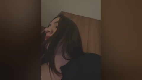 Media: Video of a woman with long, dark hair, wearing a black shirt, peeking through a wooden door, tongue sticking out, blurred background.