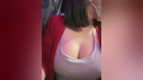 Media: Video of a woman with medium skin tone, shoulder-length black hair, wearing a low-cut white tank top and red cardigan, revealing ample cleavage, against a blurred background with red and blue hues.