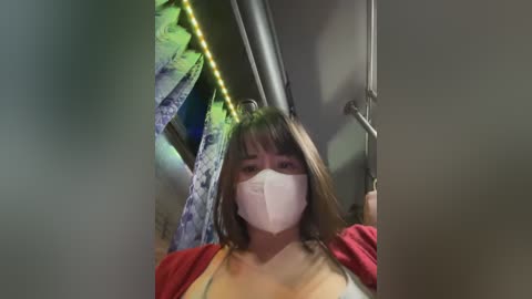 Media: Video of a woman with shoulder-length brown hair and a white face mask, wearing a red cardigan over a yellow top, holding a pole in a dimly lit room with green string lights.