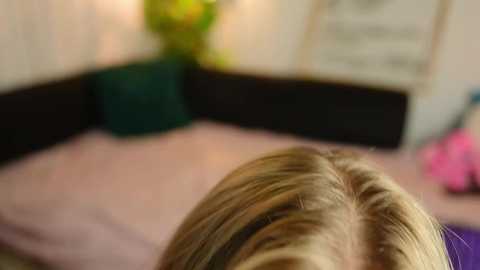 A video of a blonde woman's back and shoulder, in a cozy room with a pink bed, green plant, and blurred background.