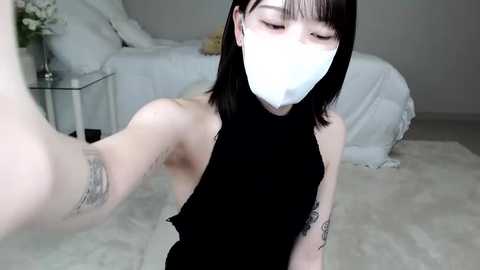 Media: A video of a pale-skinned woman with black hair, wearing a black sleeveless top, a white mask, and tattoos on her arms, taking a selfie in a minimalist bedroom with a white bed and light-colored walls.