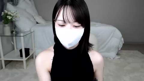 Media: A video of an Asian woman with short black hair, wearing a white mask and a black sleeveless top, sitting on a white rug in a minimalist, light-colored bedroom with a bed, side table, and vase.