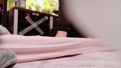 Video of a person lying on a bed, with light pink sheets and gray socks visible. A flat-screen TV in the background shows a green plant.