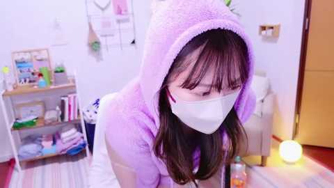 Video of a young Asian woman with long brown hair, wearing a pink fuzzy hoodie and white face mask, bending over in a brightly lit, organized room with shelves, a white couch, and a lit lamp.