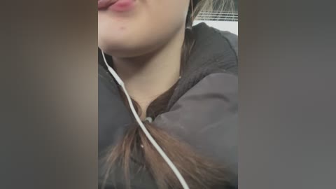 Video of a close-up of a young woman's face with light skin and long brown hair, wearing a dark hoodie and white earphones. She has a slight smile and her lips are slightly parted.