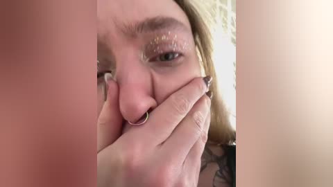 Media: A close-up video shows a woman with fair skin and blonde hair, wearing glitter eyeshadow and a nose ring, covering her mouth with her hand.