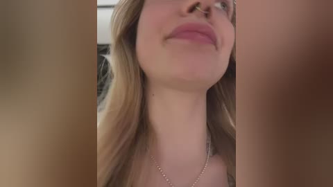 Video of a smiling young woman with fair skin and light brown hair, wearing a silver necklace, taken from a low angle. The blurred background suggests an indoor setting.