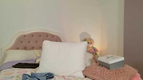 Media: Video of a cozy bedroom with a pastel-colored bed, a stuffed bunny, a soft pink pillow, a gray box, and a fluffy blanket.