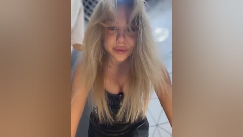 A candid video of a blonde woman with messy hair, wearing a black tank top, taken from a low angle, emphasizing her playful expression and casual attire in a modern, tiled room.