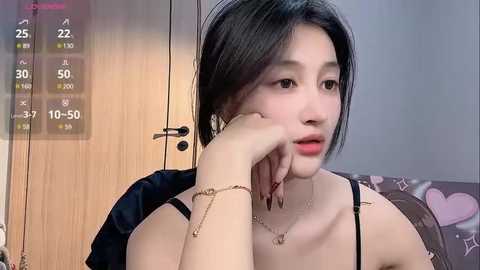 Media: Video of an Asian woman with fair skin, black hair, wearing a black spaghetti-strap top, sitting on a heart-patterned couch. She has a contemplative expression, resting her chin on her hand, and a digital thermometer displays next to her.