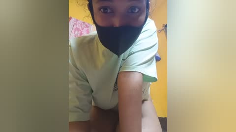 Video of a young woman with dark skin, wearing a light green T-shirt, black face mask, and no pants, crouching in a dimly lit room with yellow walls and pink patterned fabric.