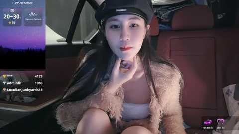 Media: Video of an Asian woman with long black hair, wearing a black cap, beige fur jacket, and white top, sitting in a car at night.