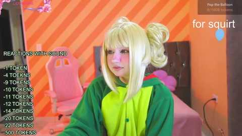 Media: Video of a pale-skinned, blonde-haired woman in a green and yellow anime cosplay, sitting at a pink gaming chair in a room with orange striped walls.