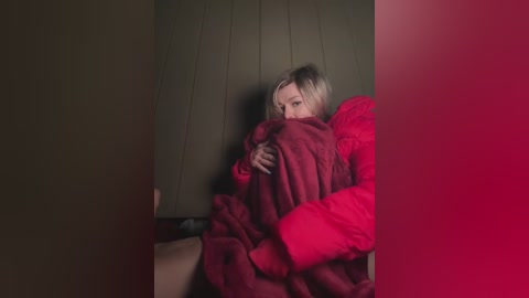 Media: Video of a blonde woman, partially covered by a red blanket, sitting on a wooden bench in a dimly lit, dark room.