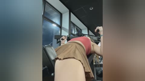 Media: Video of a woman in a gym, wearing a red tank top and brown pleated skirt, lifting weights, with blurred background.