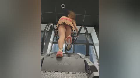 Media: Video of a woman with a blurred face, wearing a pink and brown skirt, running on an escalator in a modern, indoor setting with gray walls and a tiled ceiling.