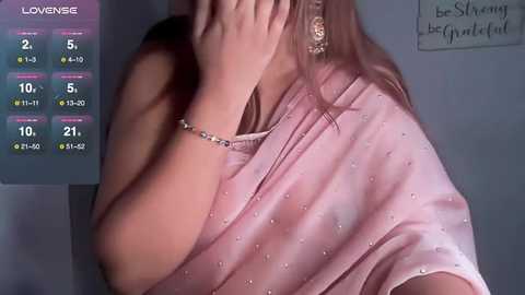 Media: Video of a woman in a pink saree with sequins, covering her face with one hand. Background shows a calendar and motivational wall text.