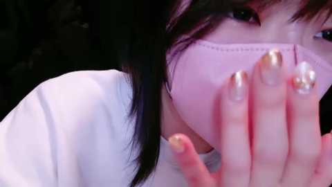 Media: Video of a woman with long black hair, wearing a white mask, with her hands covering her mouth. She has light skin and gold nail polish.