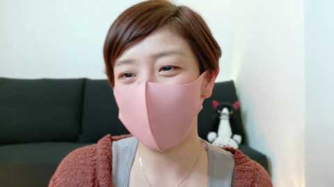 Media: Video of an Asian woman with short brown hair wearing a pink face mask, grey top, and red sweater, sitting on a black couch with a white plush toy.