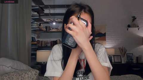 A video of a young woman with long black hair, wearing a graphic T-shirt, covering her face with a phone. Background shows a cozy, dimly-lit room with a bookshelf, plants, and a white brick wall.