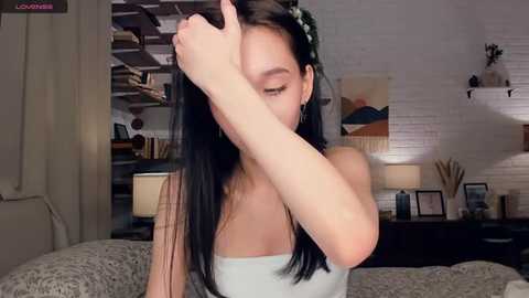 Video of an Asian woman with long black hair, wearing a white top, covering her face with her hand in a dimly lit bedroom with exposed brick walls and minimalist decor.