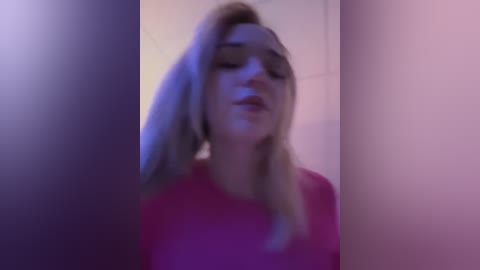 Media: Video of a young, fair-skinned woman with long blonde hair and dark sunglasses, wearing a pink top, standing in a dimly lit room with purple and pink lighting.