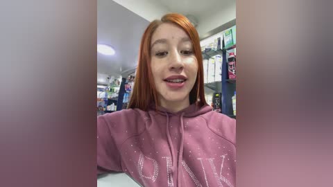 Video of a fair-skinned, red-haired young woman with a confident smile, wearing a pink hoodie, standing in a brightly lit convenience store with shelves and products visible in the background.