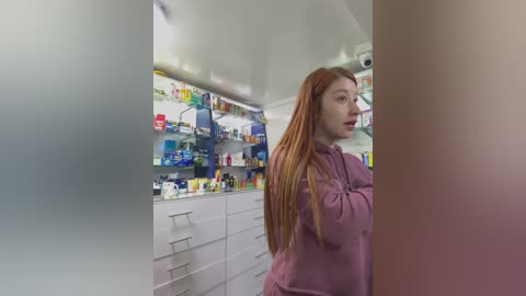 Media: Video of a young woman with long, straight red hair, wearing a purple hoodie, standing in a modern, brightly lit pharmacy. The background features shelves filled with various health products.