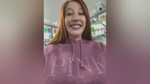 Video of a smiling Caucasian woman with long, straight red hair wearing a purple sweatshirt with \"PINK\" in white glittery letters. Background shows colorful children's books on shelves, giving a cozy, indoor reading setting.