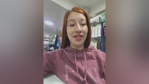 A video of a smiling redhead woman in a maroon hoodie with \"N\" on it, standing in a brightly lit drugstore aisle, surrounded by shelves filled with products.