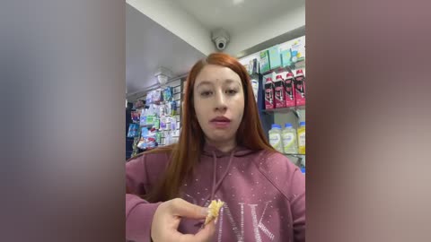 Media: A video of a young woman with long red hair, wearing a pink hoodie, holding a half-eaten pastry in a brightly lit convenience store aisle filled with shelves of snacks and drinks.