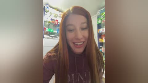 Media: Video of a smiling young woman with long red hair, wearing a maroon shirt, standing in a brightly lit kitchen with colorful food items on the shelves.