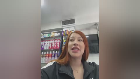 Video of a smiling young woman with long red hair, wearing a black jacket, standing in a brightly lit convenience store with shelves full of drinks and snacks in the background.