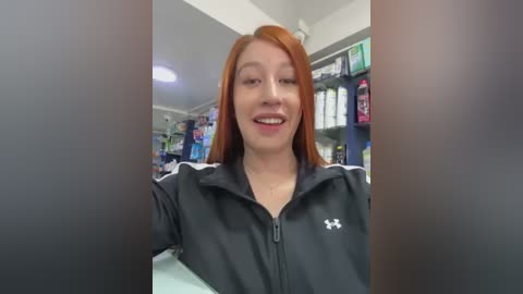 Media: Video of a young woman with long red hair, wearing a black Under Armour jacket, smiling, standing in a brightly lit store with shelves of merchandise in the background.