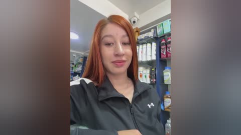 Video of a young woman with straight, shoulder-length red hair, wearing a black zip-up jacket with a white logo, standing in a well-lit pharmacy with shelves stocked with various products.