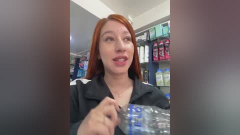 Video of a fair-skinned woman with long red hair, wearing a black jacket, holding a plastic water bottle in a modern convenience store. Shelves behind her are stocked with various beverages and snacks.