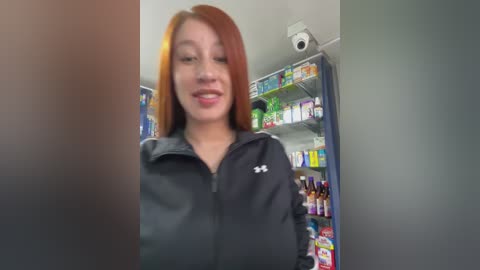 Video of a young woman with long, straight auburn hair, wearing a black zip-up jacket, standing in a brightly lit convenience store. Shelves behind her are stocked with various items.