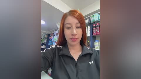 Video of a young Asian woman with straight, long red hair, wearing a black Adidas jacket, standing in a brightly lit convenience store with shelves of snacks and beverages in the background.