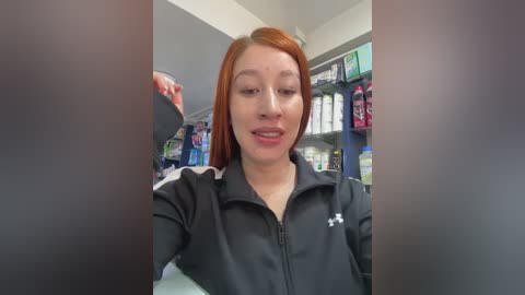 Media: Video of a young Caucasian woman with straight, shoulder-length red hair, wearing a black jacket, smiling in a pharmacy. Shelves in the background are stocked with various products.